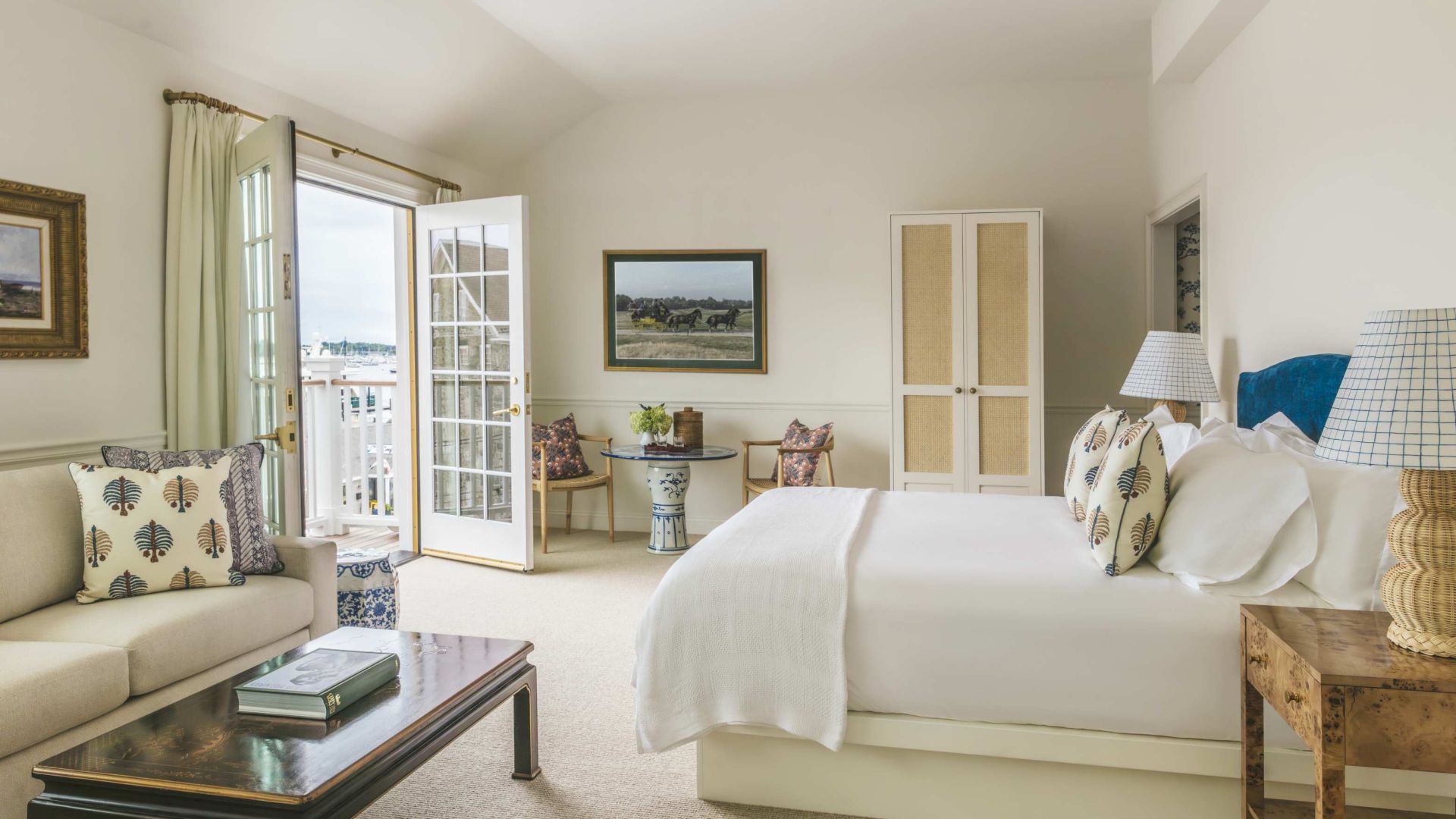 A bright, spacious Newport, RI, hotel room with wicker furniture and doors leading to a patio overlooking a serene waterfront.