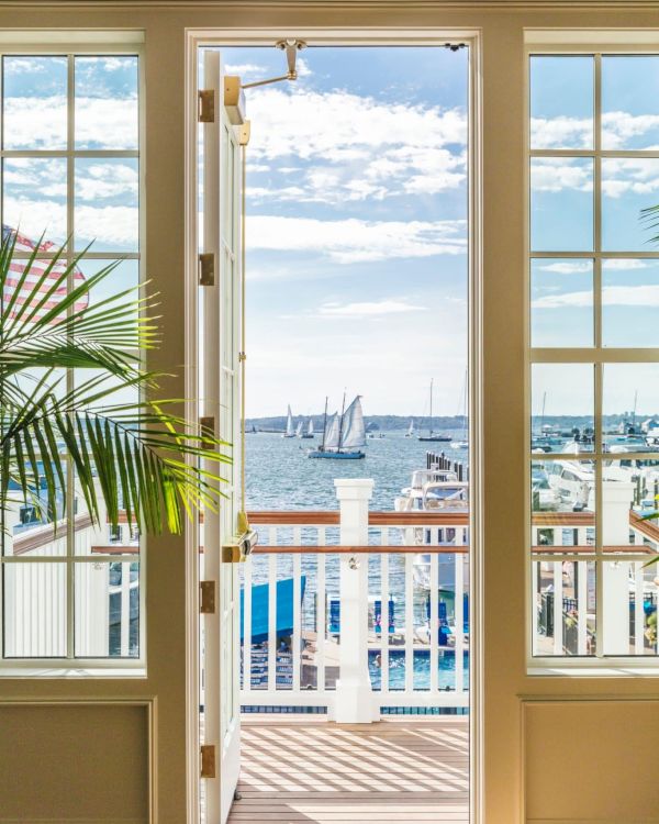 An open door leads to a balcony overlooking a scenic harbor with boats and a clear sky, framed by two large windows and a potted plant.