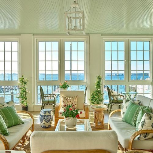 A sunlit living room with large windows, green and white decor, wicker furniture, and a view of water outside, adorned with plants and cushions.