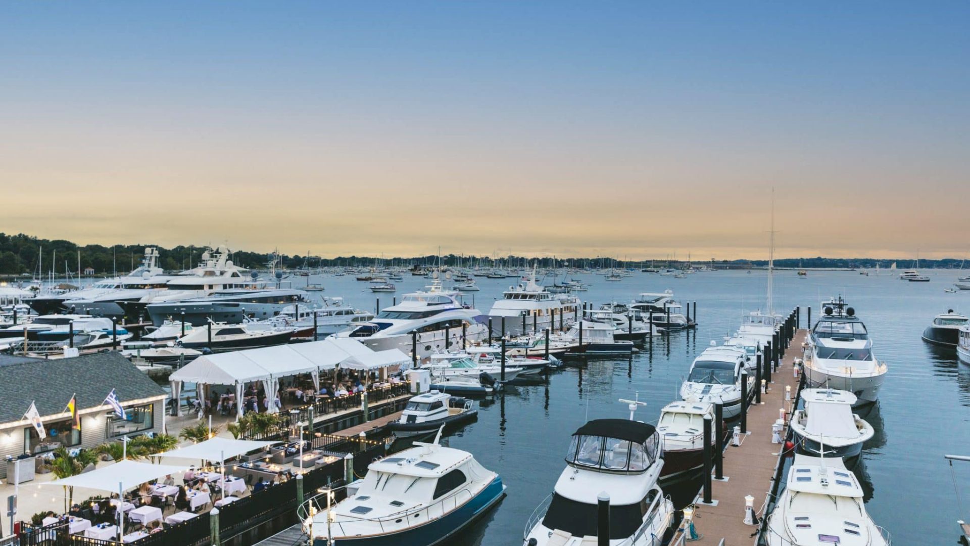 A marina at sunset filled with numerous boats and yachts, a canopy tent on the dock covering a dining area.