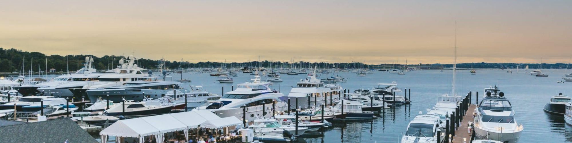 The image shows a marina filled with numerous boats and yachts, with a restaurant and dining area on the left, all set against a sunset sky.