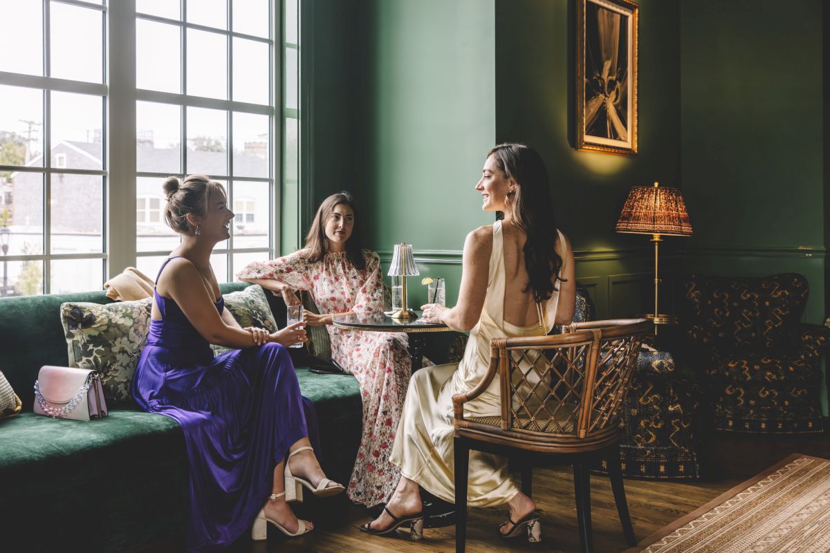 Three women sit in a lounge with green walls and a large window.