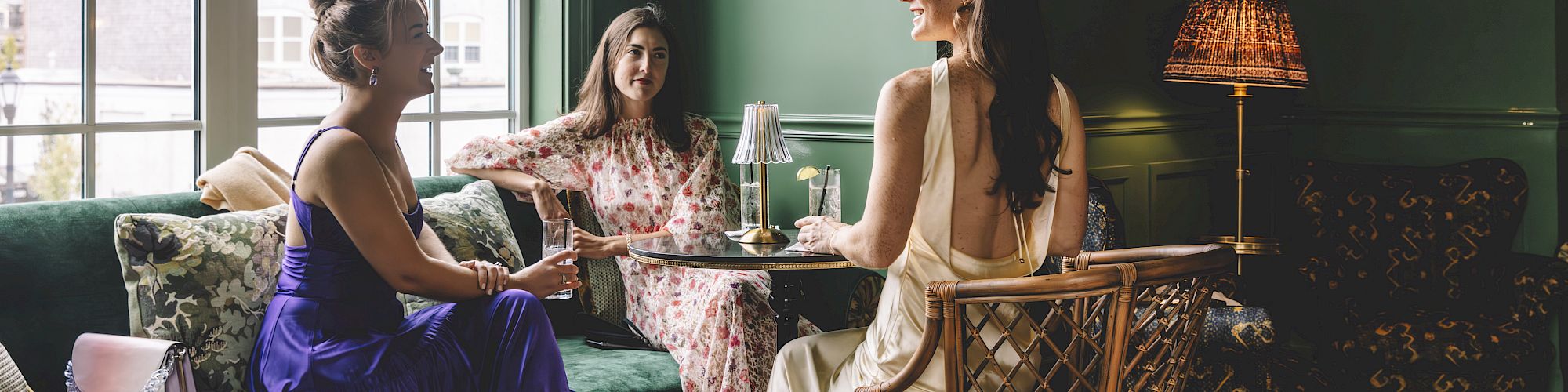 Three women are sitting in a stylishly decorated room, chatting by a table with drinks, under a unique wall decor and lamp.