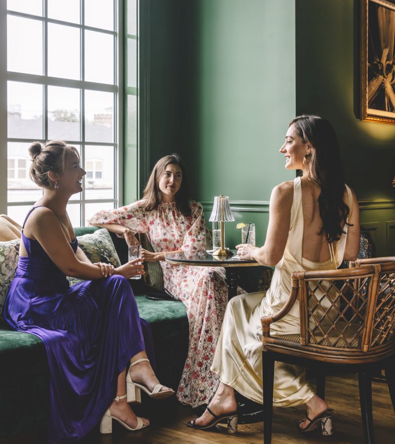 Three women in formal dresses sitting in a lobby bar with green walls, a velvet sofa, and a wooden chair.