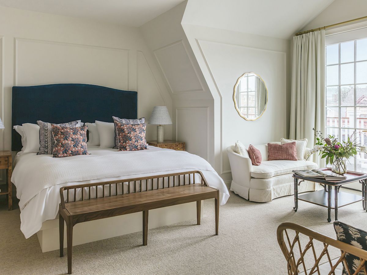 A cozy and elegant bedroom features a large bed with a blue headboard, decorative pillows, a bench at the foot, and a comfy seating area near a window.