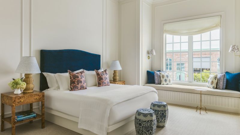 A neatly made bed with white linens and blue accents in a brightly lit room with a window seat.