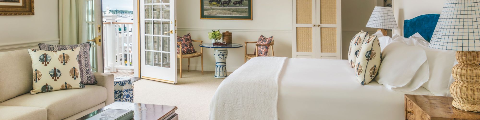 A well-decorated bedroom with a bed, a sofa, coffee table, balcony doors, chairs, lamps, and artwork on the walls, featuring neutral tones and cozy decor.