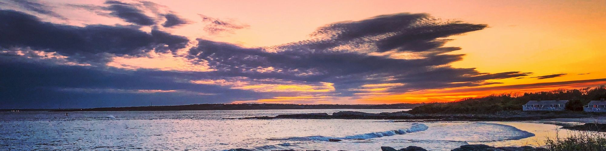 A picturesque sunset over a rocky shoreline with vibrant sky hues, gentle waves, and houses in the distance, creating a serene coastal scene.