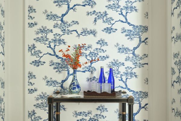 A small table with blue bottles and a white vase with flowers stands against a wall with blue and white floral wallpaper.