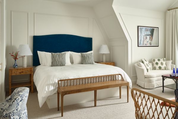 The image shows a neatly decorated bedroom with a double bed, blue headboard, a bench at the foot, side tables with lamps, and a cozy seating area.