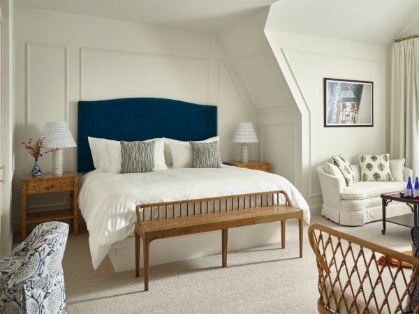 A bright, spacious Newport, RI, hotel room with a large bed, a bench, arm chairs, lamps, and a sitting area with a sofa and table.