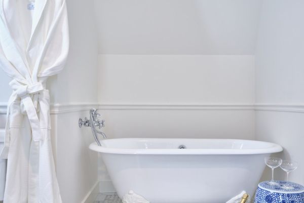 The image shows a bathroom with a white bathtub, a white bathrobe hanging, a blue and white decorative stool, and a white bath mat on a tiled floor.