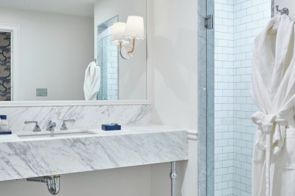 This image shows a modern bathroom with a marble countertop, large mirror, wall sconce, glass shower, and a white robe hanging on the wall.