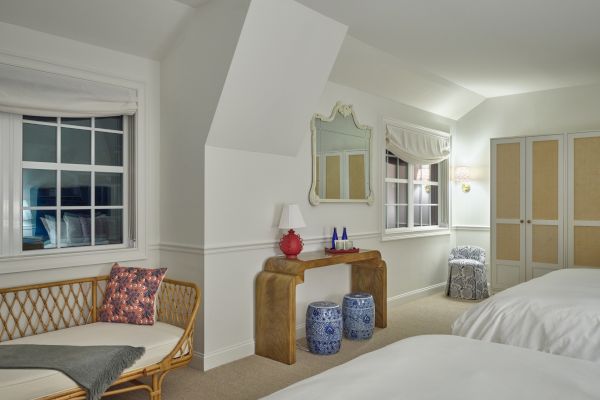 The image shows a cozy, neatly arranged bedroom with two beds, a wooden console, decorative items, two windows, a wicker bench, and a small seating area.