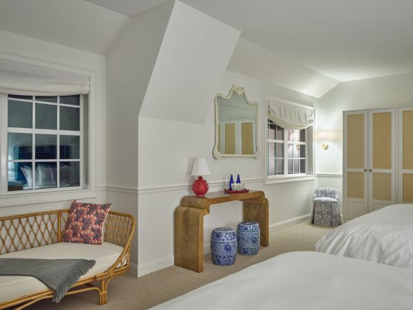A cozy Newport, RI, hotel room with two beds, a wooden console, two windows, a wicker bench, and a small seating area.