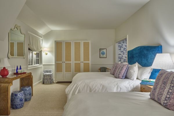 A cozy bedroom with two white beds, blue headboards, a wooden table with decor, a large mirror, and decorative stools. The sentence ends.