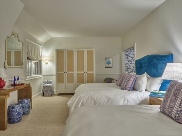 A cozy Newport, RI, hotel room with two white beds, a wooden table with decor, a large mirror, and decorative stools.