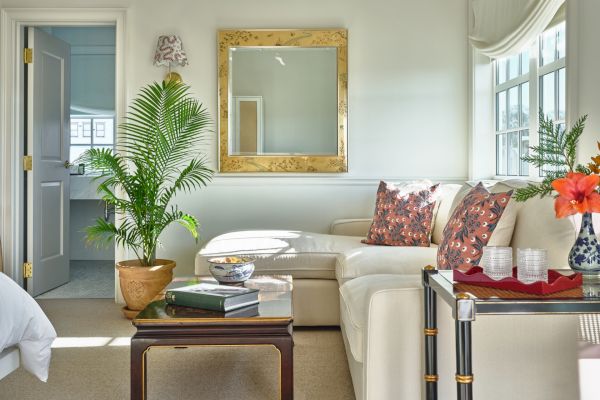 A cozy living room with a large mirror, potted plant, books on a coffee table, a cream sofa with printed pillows, and a vase with flowers.