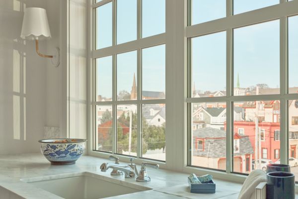 A kitchen or bathroom sink area with a marble countertop, window view of buildings, a decorative bowl, soap dispenser, tissue box, and a wall lamp.