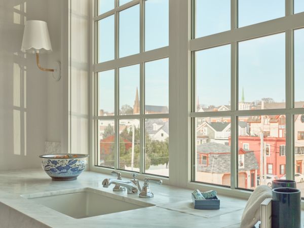 A sink area in a Newport, RI, hotel room with a marble countertop, window view of buildings, soap dispenser, and a wall lamp.