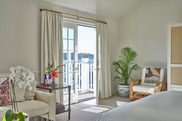 A bright room with a bed, a chair, a table with flowers, and a potted plant, featuring French doors leading to a balcony overlooking water.