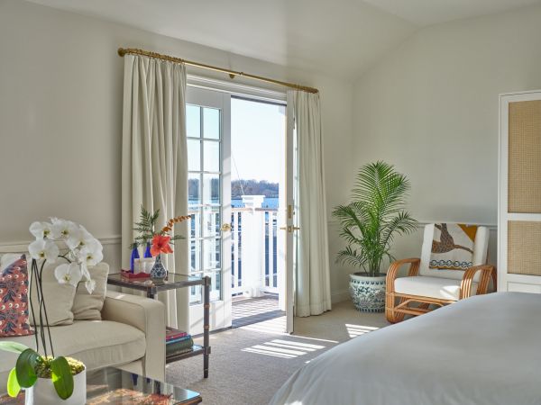 A bright Newport, RI, hotel room with a bed, an armchair, and a table featuring a balcony overlooking the Newport Harbor.