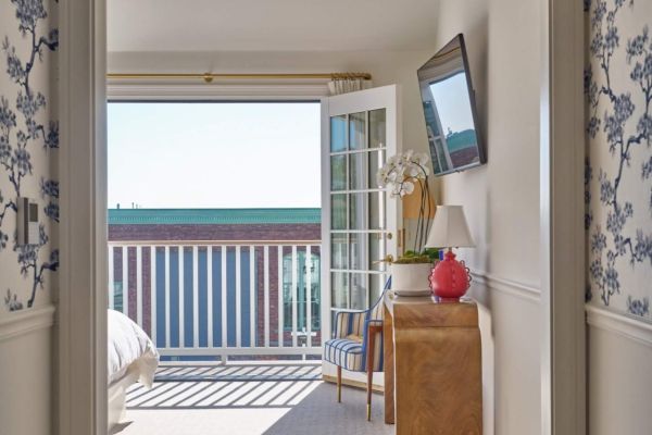 The image shows a room with floral wallpaper, a bed, a desk with a red lamp, a TV on the wall, and an open door leading to a balcony with a scenic view.