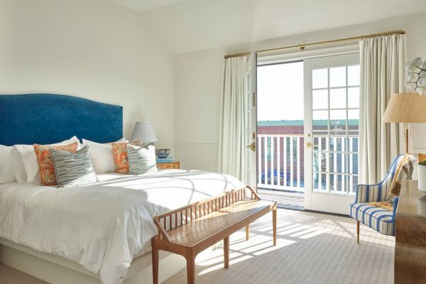 A bright bedroom with a large bed, blue headboard, bench, and striped chair. It has French doors opening to a balcony, letting in natural light.