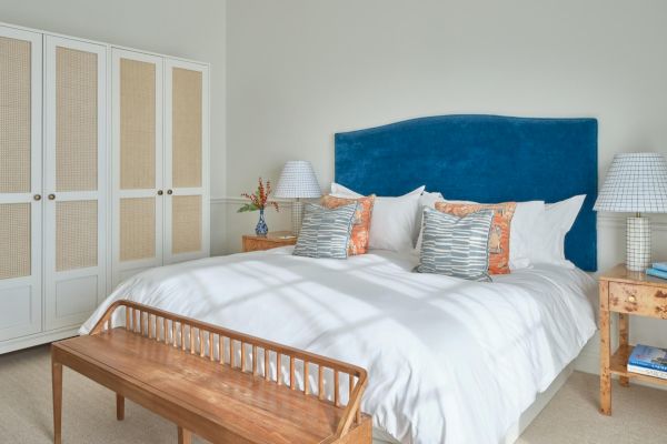 A cozy bedroom with a blue headboard, white bedspread, pillows, wooden bench, bedside tables, lamps, and a wardrobe.