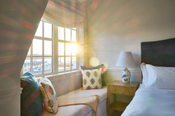 A cozy room with a bed, bedside table, and window seat featuring pillows. Sunlight streams in through the window, illuminating the space.