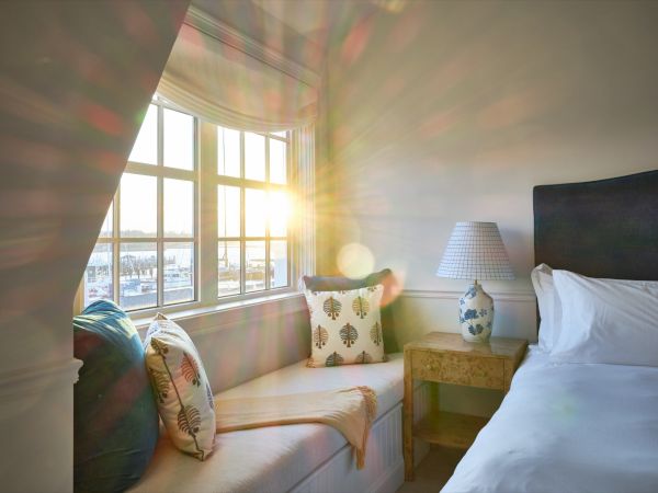 A cozy Newport, RI, hotel room with a bed, bedside table, and window seat. Sunlight streams in through the window.