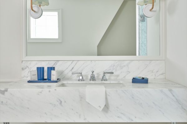 A white marble bathroom vanity with a large mirror, two sconces, and various toiletries. A small blue waste bin is on the floor.