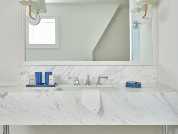 A white marble bathroom vanity in a Newport, RI, hotel room with a large mirror, two sconces, a waste bin, and various toiletries.