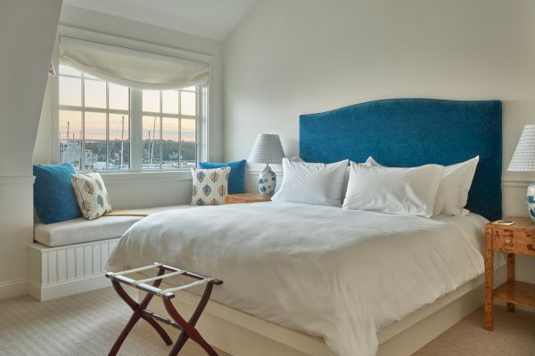 A cozy bedroom with a large white bed, blue headboard, window seat with cushions, side tables, lamps, and a scenic window view ends the sentence.