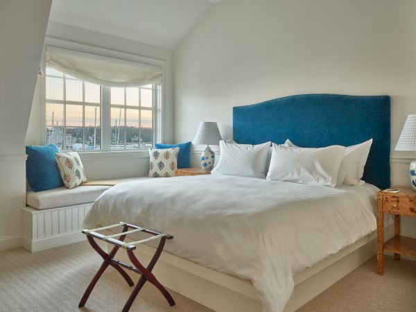 A bright Newport, RI, hotel room with a large bed, side tables, lamps, a window seat with cushions, and a scenic window view.
