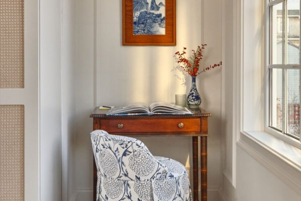 A small home office setup features a wooden desk, a floral upholstered chair, a vase with red branches, and a framed artwork on the wall.