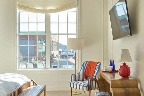 A bright bedroom with a large window, striped chair, wooden bench and desk, wall-mounted TV, and various decorative items sits within a cozy, modern space.