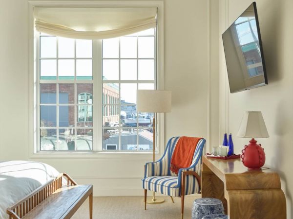 A bright Newport, RI, hotel room with a large window, striped chair, wooden bench and desk, and a wall-mounted TV.