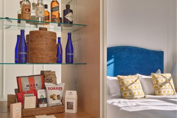 The image shows a room with a display cabinet holding bottles, glasses, and various items. In the background is a bed with pillows in yellow and blue.