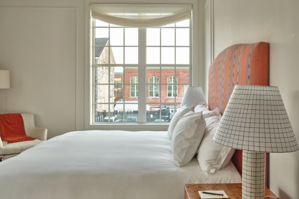 A well-lit bedroom with a large window, featuring a neatly made bed, two bedside lamps with checkered shades, and a cozy chair with an orange throw.