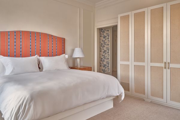 A neatly made bed with white linens and a striped headboard, bedside table with a lamp, and a white closet in a well-lit bedroom.