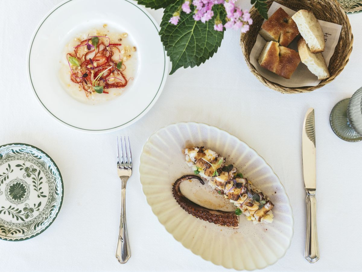 The image shows a fine dining table setting with plated gourmet dishes, a basket of bread, and floral decor on a white tablecloth.