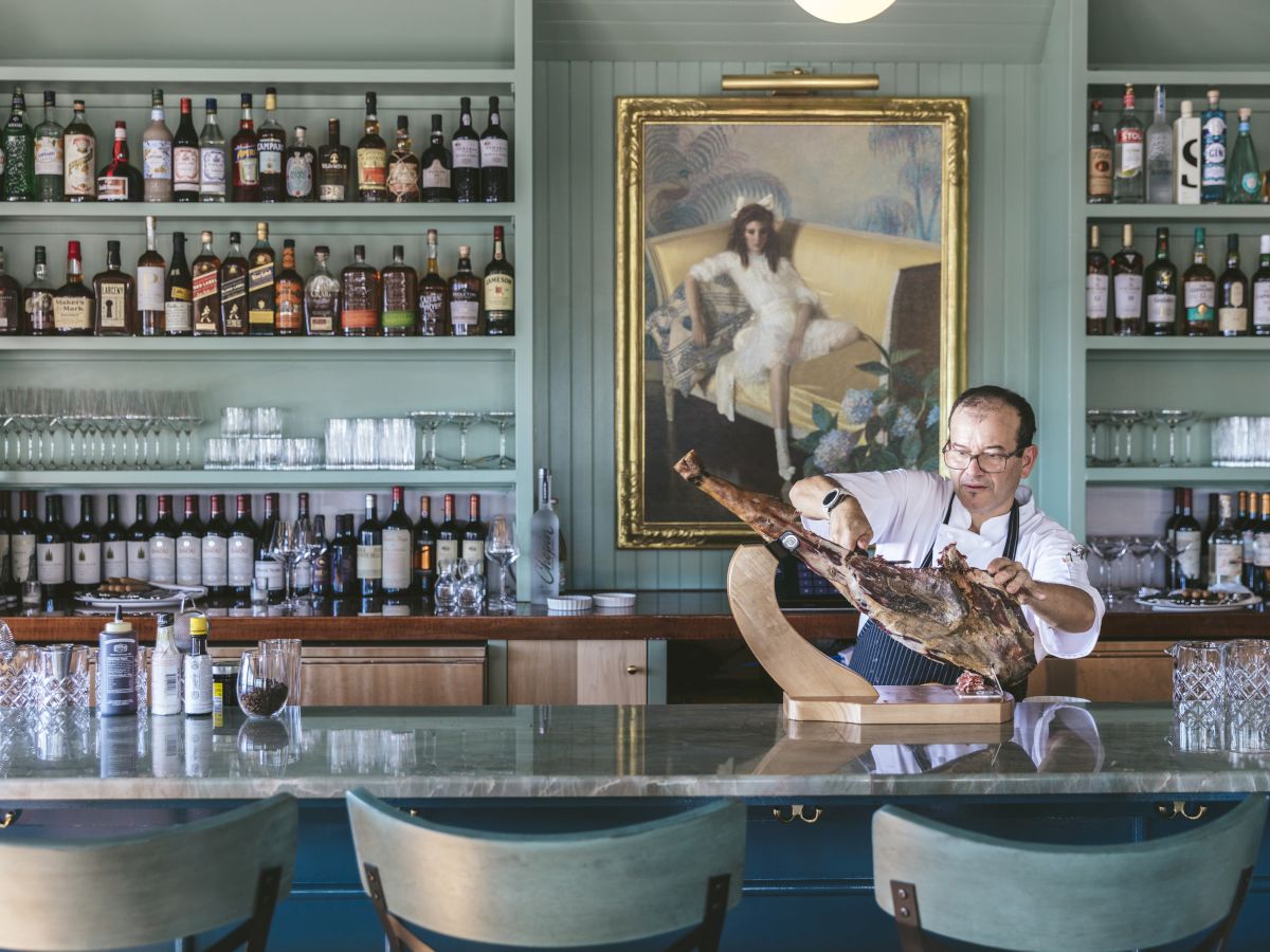 A person is cutting a piece of meat at a bar with shelves of liquor bottles, glasses, and a painting of a dog on the wall in the background.