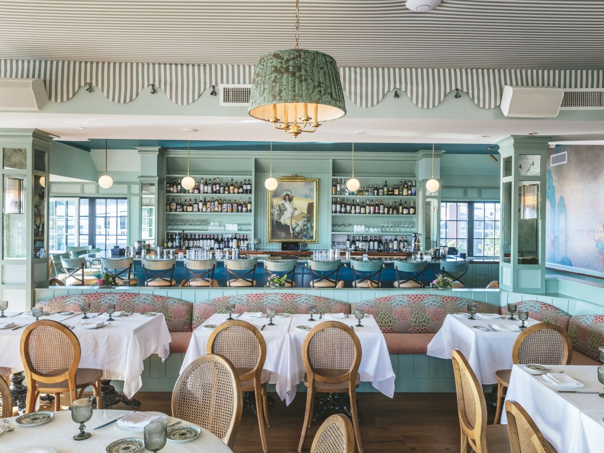 This image shows an elegant restaurant interior with tables set with white tablecloths, rattan chairs, and a bar at the back surrounded by shelves of bottles.