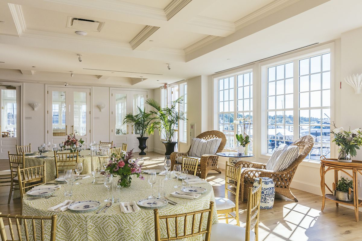 A bright event venue with round tables, chairs, elegant table settings, floral centerpieces, and large windows with harbor views.