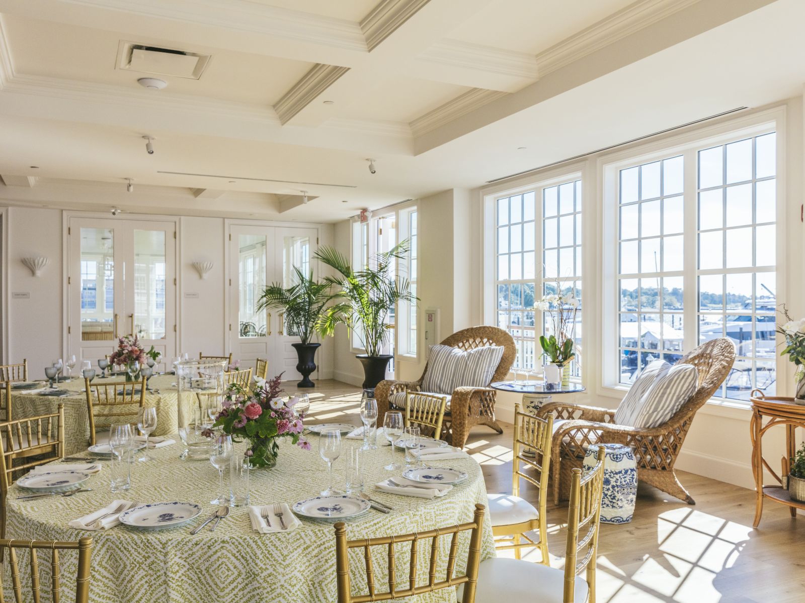 A brightly lit dining room with a round table set for dinner, gold chairs, and large windows.