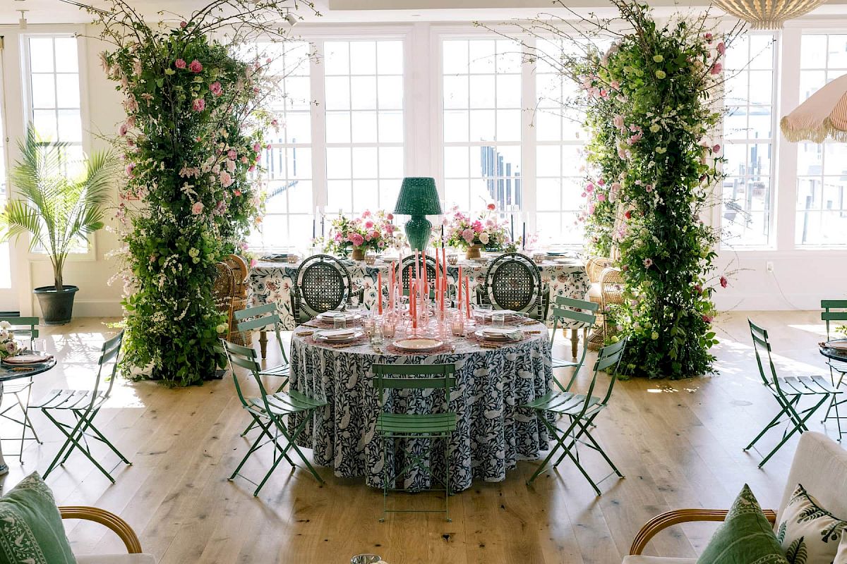 The image depicts an elegantly decorated room with a central round table, floral arrangements, greenery, and ample natural light.