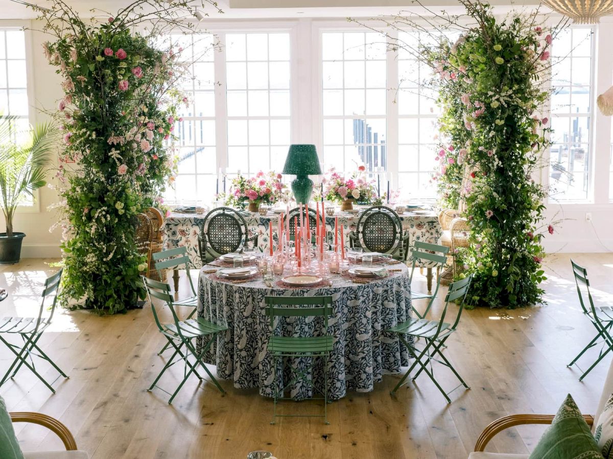 The image depicts an elegantly decorated room with a central round table, floral arrangements, greenery, and ample natural light.
