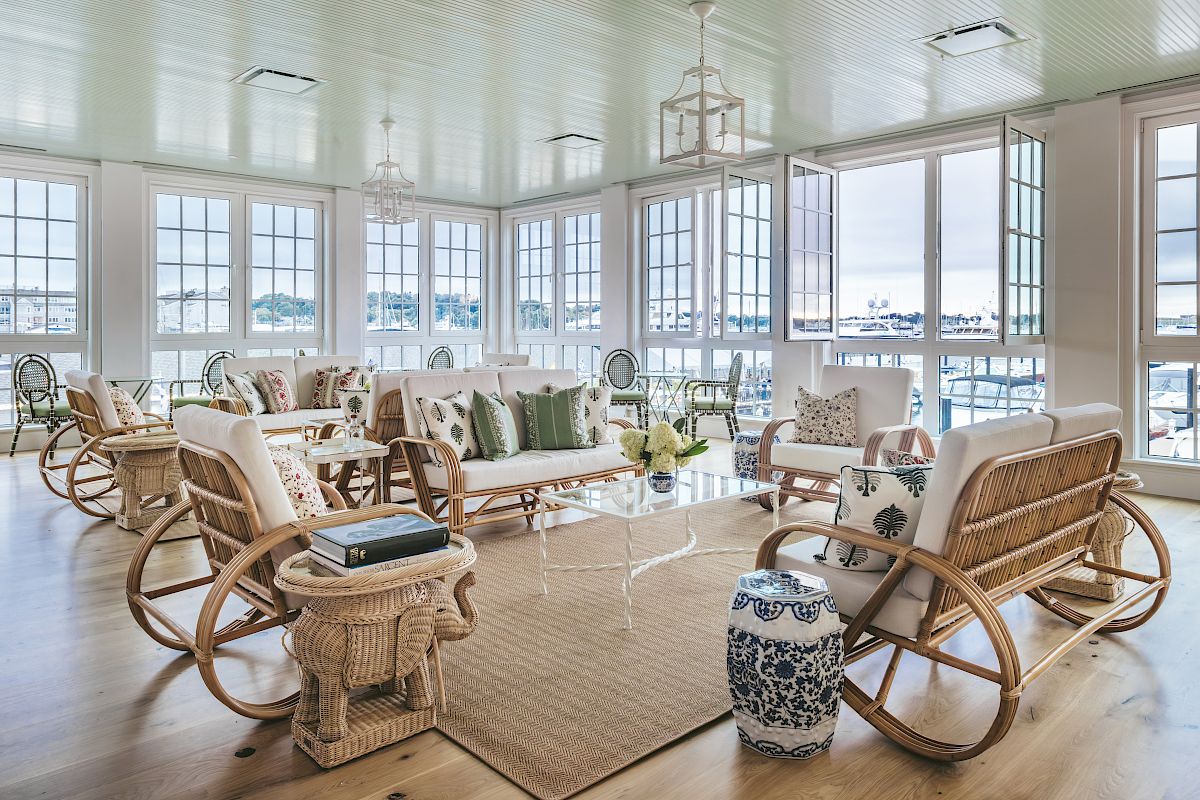A sunlit room with large windows with a view of the harbor, wicker and wood furniture with decorative pillows, and a coffee table.