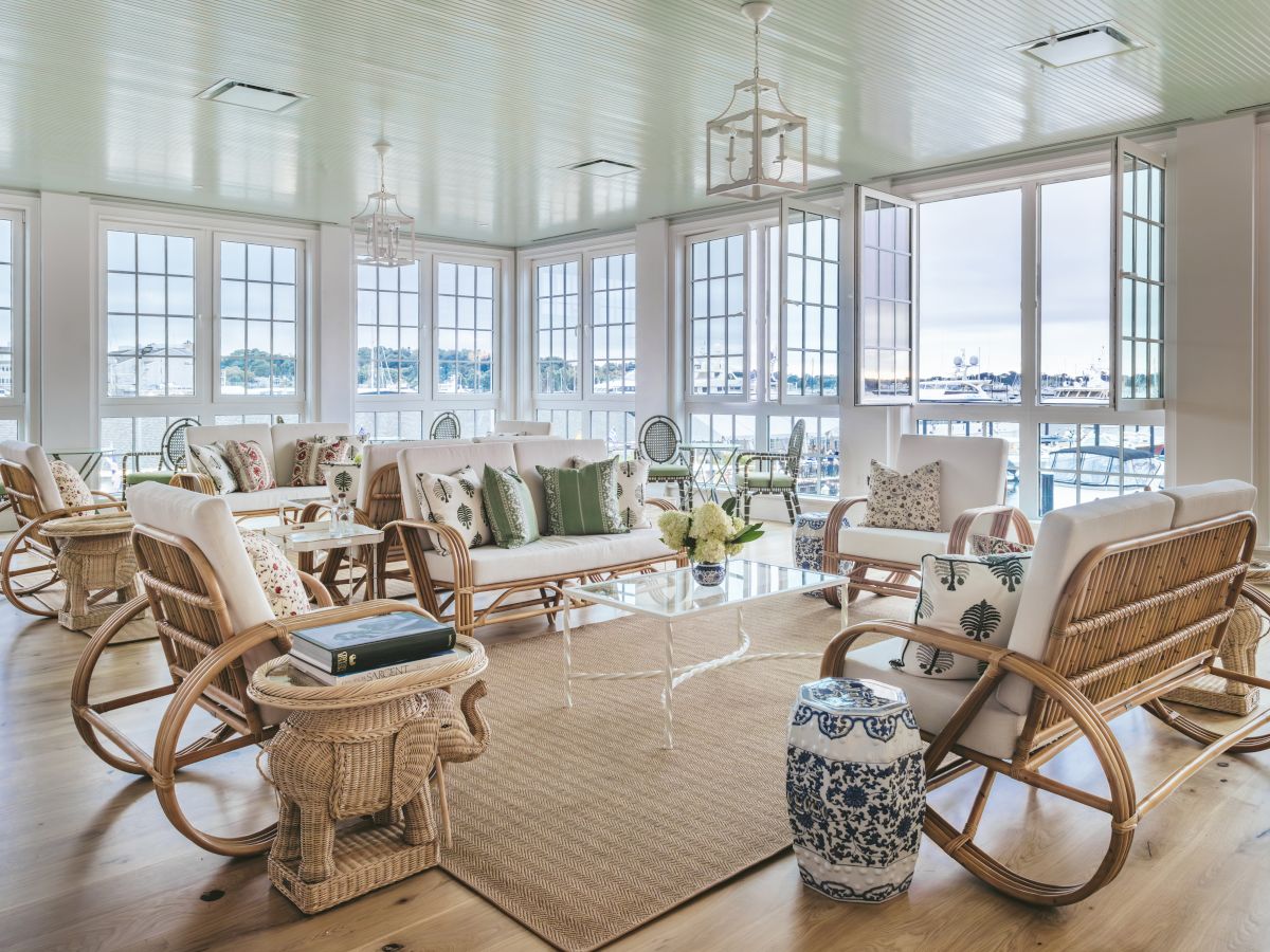 A sunlit room with large windows with a view of the harbor, wicker and wood furniture with decorative pillows, and a coffee table.